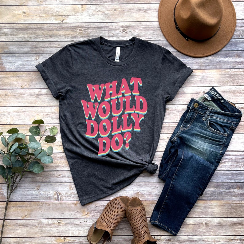 May include: A dark gray t-shirt with the text "What Would Dolly Do?" in a retro, rainbow-colored font. The shirt is laid flat on a wooden surface with a pair of brown sandals, blue jeans, and a brown hat.