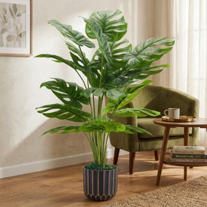 May include: A tall artificial monstera plant with large, deeply lobed green leaves. The plant is potted in a cylindrical dark navy blue planter featuring a vertical gold-toned geometric pattern around the exterior base of the container.
