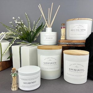 May include: A collection of white candles with wooden lids and labels. The candles are labeled "Guest House", "Luxury Hotel", and "White Tea - Thyme". There is also a white diffuser labeled "Ocean Mist" and a small glass bottle with matches.