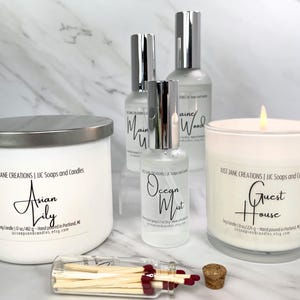 May include: Four white candles with silver lids and labels. The candles are labeled "Asian Lily", "Ocean Mist", "Guest House", and "Maine Wood". A small glass bottle of matches is in the foreground.