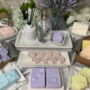 May include: Assortment of handmade soaps with bee and honeycomb designs. The soaps are displayed on white and marble trays. There are also lavender-scented candles and a diffuser.