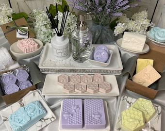 May include: An assortment of handmade soaps and bath products, including lavender-scented items. The image features soaps in various shapes, including honeycomb and floral designs, along with a reed diffuser and a soap dispenser. The color palette includes shades of purple, pink, and blue.