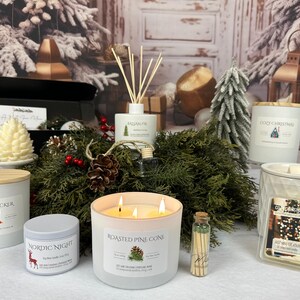 May include: A holiday-themed gift set featuring a three-wick candle labeled Roasted Pine Cone, a Nutcracker candle, a Nordic Night tin candle, a Cozy Christmas candle, a Balsam Fir reed diffuser, a wax warmer, a pine cone candle, and matches.
