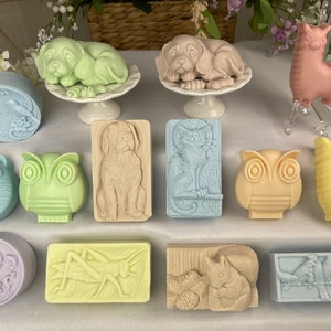 May include: A collection of artisan soaps in various shapes and colors. The soaps include animal shapes like puppies, cats, owls, and llamas, as well as floral and insect designs. The soaps are in pastel colors, including green, blue, pink, and yellow.
