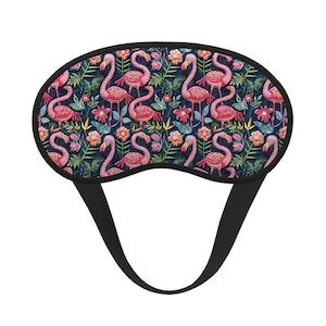 May include: A sleep mask with a black border and strap. The mask features a repeating pattern of pink flamingos, pink flowers, and green leaves on a dark blue background. The mask is designed to block out light.