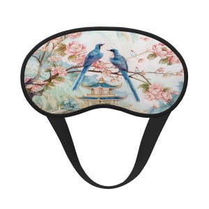 May include: A sleep mask featuring a watercolor design of two blue birds perched on a flowering branch with a pagoda in the background. The mask has a black border and a black elastic strap for a comfortable fit.