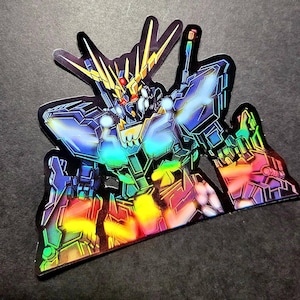 May include: Holographic sticker featuring a detailed illustration of a robot with a gold head and accents. The robot has a blue and black body with colorful rainbow highlights. The sticker is on a dark gray surface.