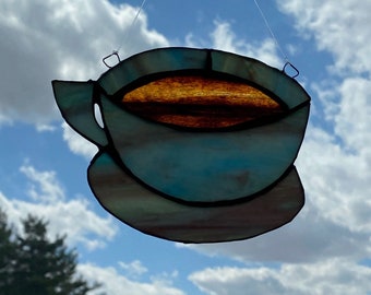 May include: A stained glass coffee cup with a blue and brown design hanging from a string against a cloudy sky.