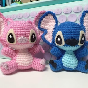 May include: Two handmade crochet dolls, one pink and one blue, resembling cartoon characters. The pink doll has large ears and a light pink belly. The blue doll has large ears and a light blue belly. Both dolls have large black eyes.