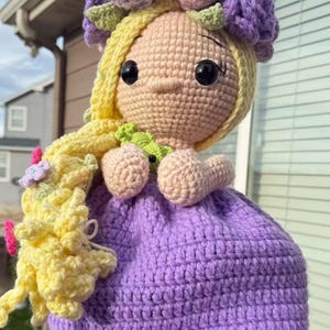 May include: A handmade crochet doll with a light skin tone, wearing a purple dress and a floral crown. The doll has yellow hair, large black eyes, and small arms. The crown features pink, purple, and green flowers.