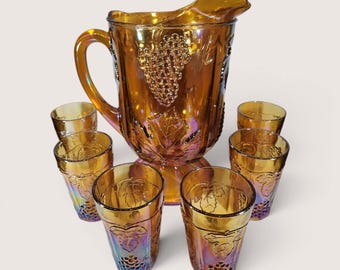 May include: An iridescent amber glass pitcher and tumbler set. The pitcher features a handle and a grape design. The set includes six matching tumblers, also with a grape design. The glassware has a vintage aesthetic.