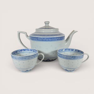 May include: A white porcelain tea set with blue decorative trim. The set includes a teapot with a lid and two matching teacups. The cups and teapot have a delicate, dotted pattern. The tea set is displayed on a plain background.