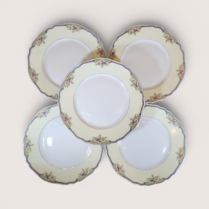 May include: Five cream-colored plates with scalloped edges. Each plate features a floral design in pink, yellow, and green around the rim, with a navy blue border and a gold trim. The center of each plate is white.