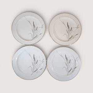 May include: Four white ceramic plates with a wheat stalk design in gray and gold trim. The plates are arranged in a square, showcasing the elegant, minimalist design. The plates are suitable for serving meals.