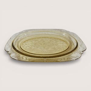 May include: A vintage, oval-shaped amber glass serving tray. The tray features an ornate, embossed floral design in the center and along the scalloped edges. The tray is a light amber color and is likely made of pressed glass.