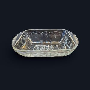 May include: Clear glass serving dish with an oval shape and decorative floral and leaf patterns. The dish has a scalloped rim and is designed with embossed details. The glass is transparent, allowing light to pass through, and is suitable for serving food.