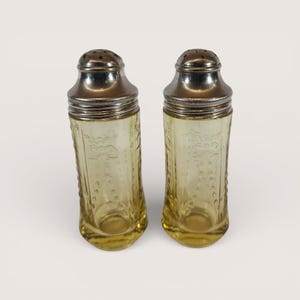 May include: A pair of vintage salt and pepper shakers. The shakers have a clear, yellow-tinted glass body with an embossed floral design. They are topped with a silver-colored metal lid with small holes for dispensing. The shakers have a curved, hourglass shape.