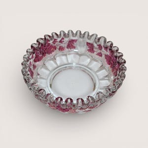 May include: A clear glass bowl with a ruffled edge and a circular base. The bowl features a delicate pattern of maroon-colored leaves and vines around the rim. The bowl is designed with a scalloped edge.