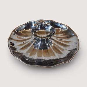 May include: A silver-colored, scalloped serving dish with a central, smaller bowl. The dish has a reflective surface and a fluted design radiating from the center. The overall shape resembles a seashell.