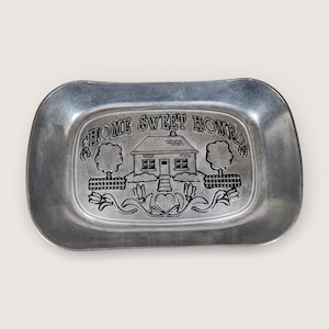 May include: A rectangular, pewter-colored tray with rounded corners. The tray features an embossed design with the words "HOME SWEET HOME" above a house illustration, trees, and floral elements. The tray is likely used for decorative purposes.