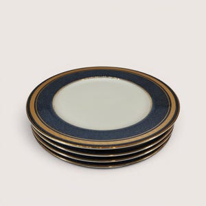 May include: A stack of four white ceramic plates with a navy blue border and a gold band. The plates have a simple, elegant design, suitable for formal dining or special occasions. The plates are stacked neatly on a white surface.