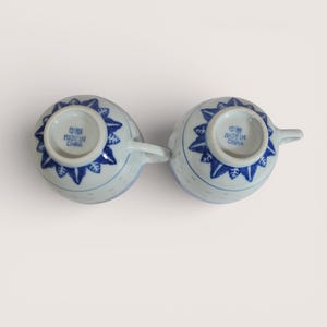 May include: Two white ceramic teacups with blue floral designs and handles. The cups are upside down, revealing the blue floral pattern on the bottom. The text "MADE IN CHINA" is visible on the bottom of each cup.