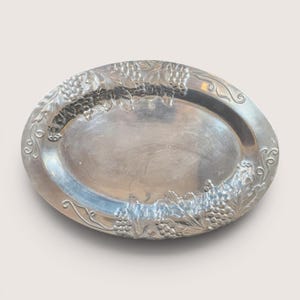 May include: Oval silver-toned serving tray with embossed grape and leaf designs around the rim. The tray has a polished, reflective surface and is likely made of metal. A decorative piece for serving food or displaying items.