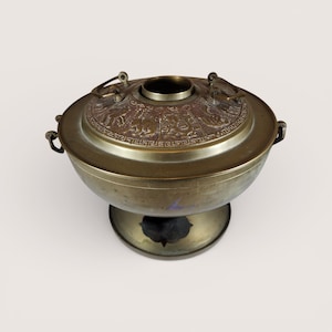 May include: A vintage brass vessel with a lid featuring an embossed design. The vessel has a round body, a pedestal base, and handles on the sides. The lid has a central opening and decorative elements.