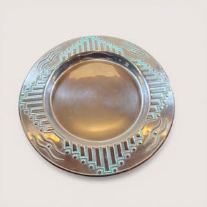 May include: A silver-colored plate with a decorative border. The border features a geometric design with teal accents. The plate has a slightly raised edge and a smooth, reflective surface. The plate is round and appears to be made of metal.