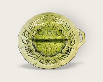 May include: A vintage green glass divided serving dish with ornate floral and geometric patterns. The dish has a round shape with a handle on one side and is divided into two sections. The glass has a slight yellow tint.