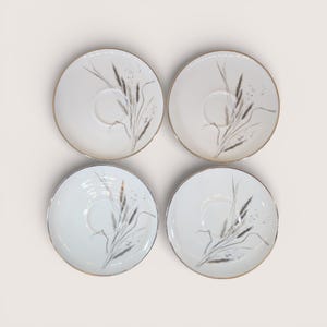 May include: Four white ceramic saucers with a gold rim and a delicate gray wheat stalk design. The saucers are arranged in a square, showcasing their elegant, minimalist aesthetic. Ideal for a tea set or a vintage collection.