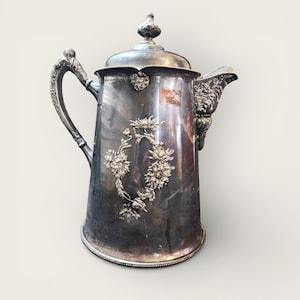 May include: A silver-colored antique coffee pot with ornate floral detailing. The pot features a curved handle, a spout, and a decorative lid with a finial. The body of the pot is adorned with a floral design, adding to its vintage charm.