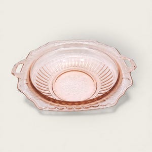 May include: A pink glass serving bowl with a scalloped edge and two handles. The bowl has a ribbed interior and a floral design in the center. The dish is likely vintage and suitable for serving food.