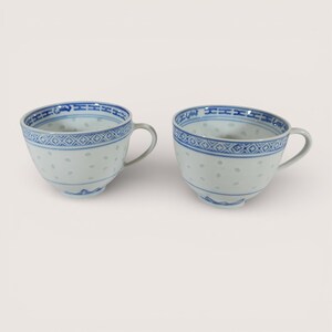 May include: Two white porcelain teacups with blue decorative trim and handles. The cups have a delicate, dotted pattern and a detailed blue border around the rim. The design is repeated on both cups, suggesting a matching set.