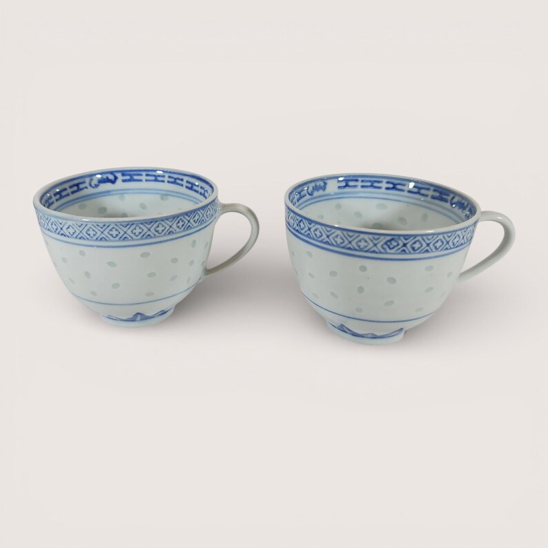May include: Two white porcelain teacups with blue decorative trim and handles. The cups have a delicate, dotted pattern and a detailed blue border around the rim. The design is repeated on both cups, suggesting a matching set.