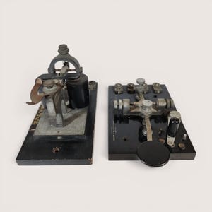 May include: Two vintage telegraph keys mounted on black rectangular bases. The left device features a metal arm and a black cylindrical coil. The right device includes a black circular knob, metal adjustment screws, and text reading J.H. BUNNELL & CO.