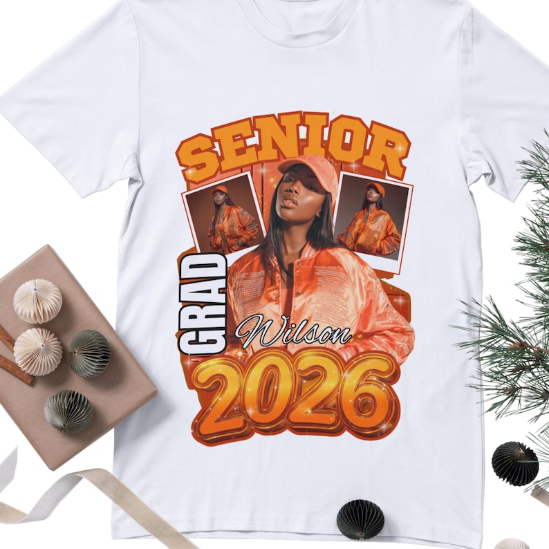 May include: White t-shirt featuring a graduation design. The design includes the word "SENIOR" in orange, the word "GRAD" in white, and the name "Wilson" with the year "2026". The design also features a photo of a person wearing an orange jacket and cap.
