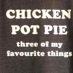 May include: Dark gray t-shirt with the words "CHICKEN POT PIE" in large, white, bold letters. Below, the text reads "three of my favourite things" in a smaller, white font.