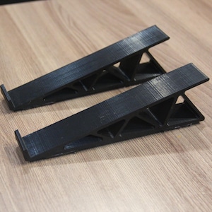 May include: Two black, angled laptop stands are displayed on a light brown wooden surface. The stands have a triangular support structure and a small lip to hold a device. The stands are designed to elevate a laptop.