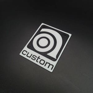 Può includere: Adesivo in plastica bianca con un logo circolare nero e la parola "custom" stampata sotto.