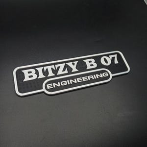 以下が含まれることがあります： 白黒の3Dプリントされたサインで、白い文字で「Bitzy B 07 Engineering」と書かれています。