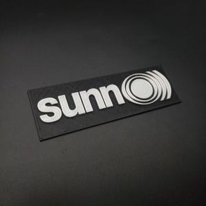 May include: A black rectangular label with the word "sunn" in white raised lettering and a white stylized circle design.