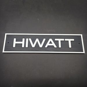 Puede incluir: Un letrero de madera negro con un borde blanco y la palabra "HIWATT" en letras blancas.