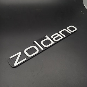 Puede incluir: Emblema de plástico blanco con la palabra "zoldano" en letras en relieve.