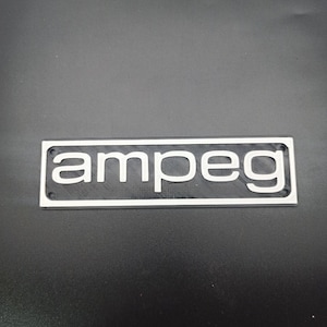 May include: A silver and black rectangular plate with the word "Ampeg" printed in white letters.