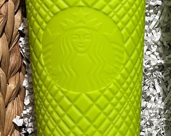 Op de afbeelding: Een felgroene herbruikbare Starbucks-beker met een ruitpatroon en het Starbucks-logo. De beker staat in een mand gevuld met wit gescheurd papier. De tekst op de bodem van de beker luidt "Starbucks, Aka.A Distribuidora e Importadora S.A. de C.V., Carretera Lago de Guadalupe km 17.7, Ext. SN, Nave 8, San Bartolo, Tlalnepantla, Edo. de México, México, C.P. 54010, RFC DIAP10808STE, HECHO EN CHINA."
