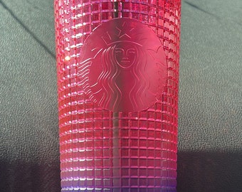 Op de afbeelding: Een roze en blauwe ombre Starbucks-beker met een rasterpatroon en het Starbucks-logo. De beker heeft een groen deksel en een rietje.