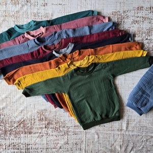 Könnte beinhalten: Ein Stapel von 12 langärmeligen Waffelstrick-Shirts in verschiedenen Farben, darunter Grün, Gelb, Orange, Rot, Blau und Lila. Die Shirts sind fächerförmig angeordnet, wobei das grüne Shirt unten liegt.