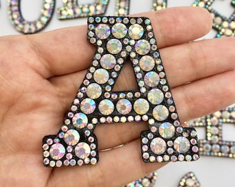 Può includere: Una grande lettera A scintillante e iridescente realizzata con strass su uno sfondo in feltro nero. La lettera è tenuta in una mano.