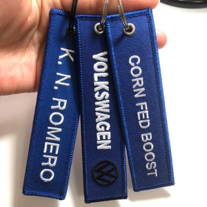 Può includere: Tre portachiavi blu con testo bianco. I portachiavi recitano "K.N. Romero", "VOLKSWAGEN" con un logo Volkswagen e "CORN FED BOOST".